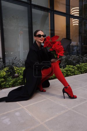 Téléchargez les photos : Photo de mode en plein air de belle femme brune en robe élégante, manteau noir et accessoires, marche dans la rue avec beau bouquet de fleurs rouges poinsettia - en image libre de droit