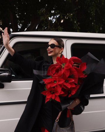 Foto de Foto de moda al aire libre de la hermosa mujer morena en vestido elegante, abrigo negro y accesorios, caminando por la calle con hermoso ramo de flores rojas de poinsettia - Imagen libre de derechos