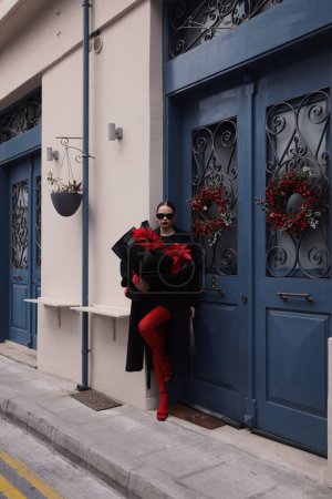 Téléchargez les photos : Photo de mode en plein air de belle femme brune en robe élégante, manteau noir et accessoires, marche dans la rue avec beau bouquet de fleurs rouges poinsettia - en image libre de droit