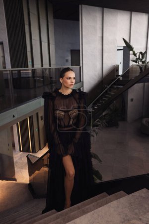 Téléchargez les photos : Photo de mode de belle femme avec hai foncé dans la robe noire élégante posant dans le hall de l'hôtel de luxe - en image libre de droit