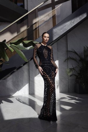 Téléchargez les photos : Photo de mode de belle femme aux cheveux foncés dans une élégante robe noire avec des accessoires posant dans le hall de l'hôtel de luxe - en image libre de droit