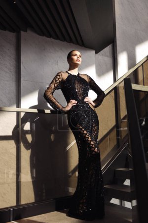 Téléchargez les photos : Photo de mode de belle femme aux cheveux foncés dans une élégante robe noire avec des accessoires posant dans le hall de l'hôtel de luxe - en image libre de droit