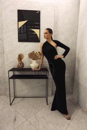 Foto de Foto de moda de una hermosa mujer con cabello oscuro en elegante vestido negro con accesorios posando en el lujoso vestíbulo del hotel - Imagen libre de derechos