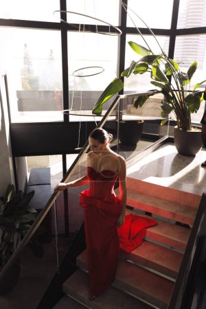 Téléchargez les photos : Photo de mode de belle femme aux cheveux foncés dans une élégante robe rouge avec des accessoires posant dans le hall de l'hôtel de luxe - en image libre de droit