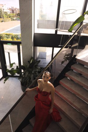 Téléchargez les photos : Photo de mode de belle femme aux cheveux foncés dans une élégante robe rouge avec des accessoires posant dans le hall de l'hôtel de luxe - en image libre de droit