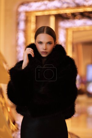 Foto de Foto interior de moda de hermosa mujer sensual con cabello oscuro en lujoso abrigo de piel negro y joyas posando en el vestíbulo del hotel - Imagen libre de derechos