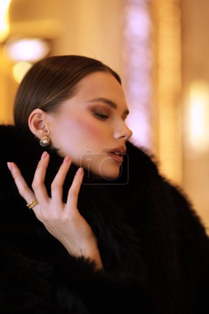 Téléchargez les photos : Photo d'intérieur de mode de belle femme sensuelle aux cheveux foncés en manteau de fourrure noir luxueux et bijoux posant dans le hall de l'hôtel - en image libre de droit