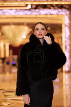 Foto de Foto interior de moda de hermosa mujer sensual con cabello oscuro en lujoso abrigo de piel negro y joyas posando en el vestíbulo del hotel - Imagen libre de derechos