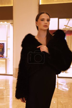 Téléchargez les photos : Photo d'intérieur de mode de belle femme sensuelle aux cheveux foncés en manteau de fourrure noir luxueux et bijoux posant dans le hall de l'hôtel - en image libre de droit