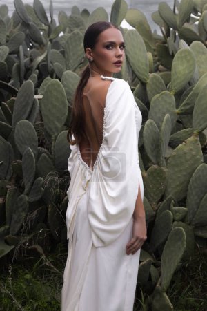 Foto de Foto de moda al aire libre de hermosa mujer con cabello oscuro en vestido de novia de lujo con joyas posando en un hermoso lugar de la naturaleza Lefkara, con plantación de cactus en el fondo - Imagen libre de derechos