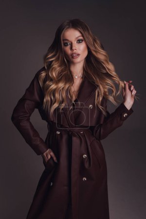 Téléchargez les photos : Photo de mode de belle femme aux cheveux blonds en cuir luxueux manteau marron posant en studio - en image libre de droit