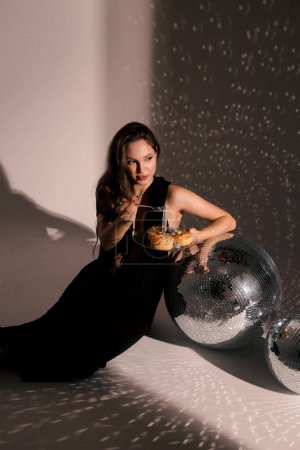 Photo for Fashion photo of beautiful pregnant woman with dark hair in elegant clothes posing in studio with disco ball, holding plate of cakes and jewelry - Royalty Free Image