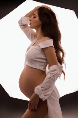 Téléchargez les photos : Photo de mode de belle femme enceinte aux cheveux foncés dans des vêtements élégants posant en studio - en image libre de droit