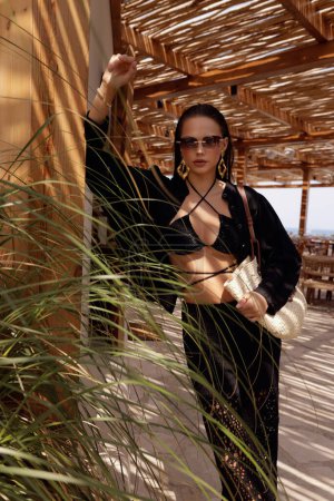 Téléchargez les photos : Photo de mode de belle femme aux cheveux foncés dans des vêtements élégants avec des accessoires posant dans le club de plage d'été - en image libre de droit