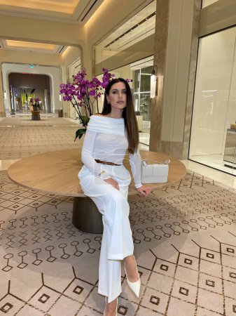 Téléchargez les photos : Photo de mode de belle femme sensuelle avec les cheveux foncés dans des vêtements blancs luxueux avec des accessoires posant dans le hall élégant hôtel - en image libre de droit
