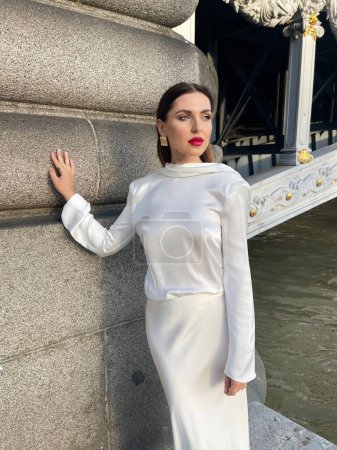 Photo for Fashion outdoor photo of beautiful woman with dark hair in luxurious white silk dress and accessories posing by the Alexandre III bridge in Paris - Royalty Free Image