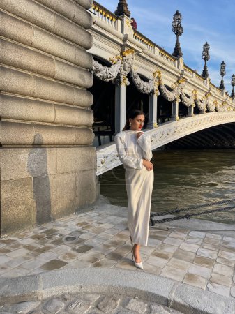 Photo for Fashion outdoor photo of beautiful woman with dark hair in luxurious white silk dress and accessories posing by the Alexandre III bridge in Paris - Royalty Free Image