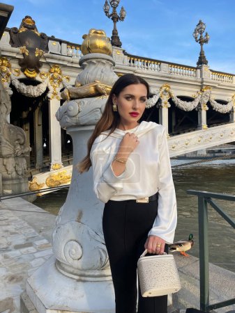 Photo for Fashion outdoor photo of beautiful woman with dark hair in luxurious white silk blouse and black pants and accessories posing by the Alexandre III bridge in Paris - Royalty Free Image