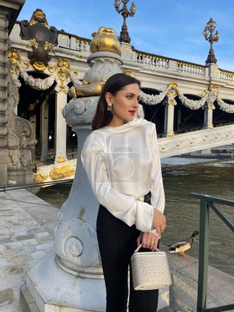 Foto de Foto de moda al aire libre de una hermosa mujer con cabello oscuro en lujosa blusa de seda blanca y pantalones negros y accesorios posando junto al puente Alexandre III en París - Imagen libre de derechos