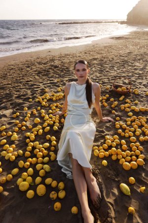 Photo for Fashion outdoor photo of beautiful woman with dark hair in elegant white dress posing on the Cyprus beach with a lot of lemons - Royalty Free Image
