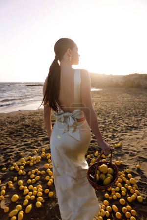 Photo for Fashion outdoor photo of beautiful woman with dark hair in elegant white dress posing on the Cyprus beach with a lot of lemons - Royalty Free Image