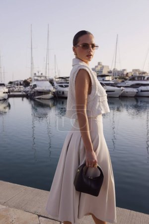 Foto de Fotos de moda al aire libre de hermosa mujer con el pelo oscuro en ropa clásica elegante con accesorios posando en el puerto deportivo con los yates en Chipre - Imagen libre de derechos