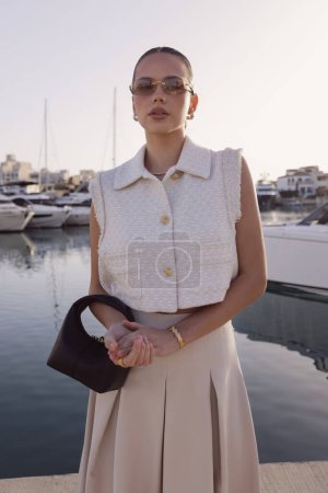 Téléchargez les photos : Photos de mode en plein air de belle femme aux cheveux foncés dans des vêtements classiques élégants avec des accessoires posant à la marina avec les yachts à Chypre - en image libre de droit