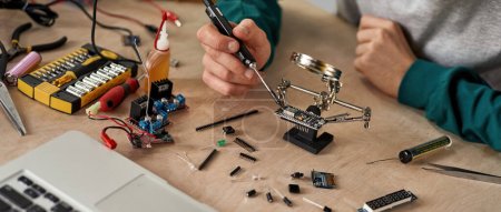 Photo for Cropped male technician or engineer soldering microchip with soldering iron under magnifying glass at table with laptop, variety technical tools and components. Modern technology and innovation - Royalty Free Image