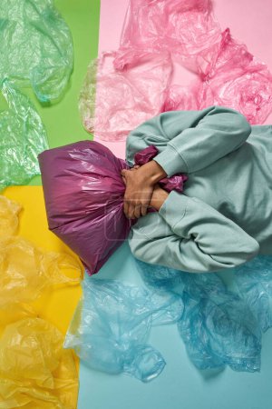 Foto de Chica negra sofocante de bolsa de plástico en la cabeza mientras está acostado sobre un fondo colorido con basura. Ecología seguridad y protección. Eliminación y reciclaje de residuos. Sostenibilidad ambiental. Sesión de estudio - Imagen libre de derechos