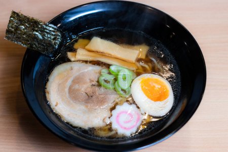 Soy sauce ramen in a black cup topped with chashu pork, Naruto fish balls, soy-sauce pickled eggs, menma bamboo shoots and seaweed. Ramen is a distinctive Japanese style noodle soup.