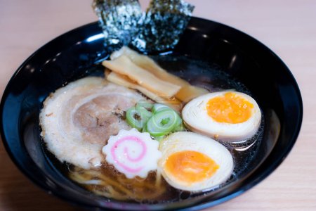 Photo for Soy sauce ramen in a black cup topped with chashu pork, Naruto fish balls, soy-sauce pickled eggs, menma bamboo shoots and seaweed. Ramen is a distinctive Japanese style noodle soup. - Royalty Free Image