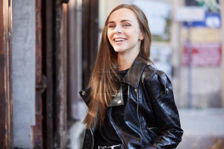 Téléchargez les photos : Belle jeune femme souriante en veste en cuir noir debout sur la rue - en image libre de droit