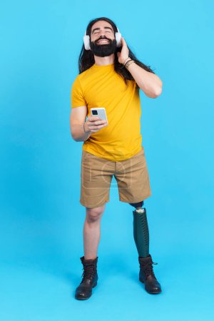 Foto de Retrato de estudio vertical con fondo azul de un hombre con pierna protésica disfrutando escuchando música con el móvil - Imagen libre de derechos
