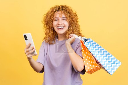 Foto de Studio image with yellow background of a young smiley woman with curly hair using the mobile while holing shopping bags - Imagen libre de derechos