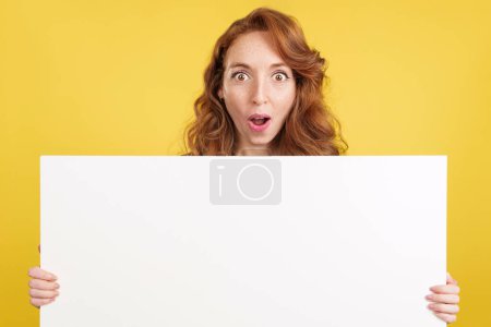 Photo for Amazed redheaded woman holding a blank panel while looking at camera in studio with yellow background - Royalty Free Image