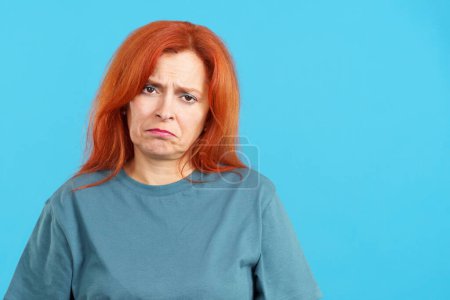 Téléchargez les photos : Femme rousse mature regardant la caméra avec une expression triste dans le studio avec fond bleu - en image libre de droit
