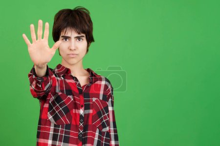 Photo for Serious androgynous person gesturing rejection with the palm of the hand in studio with green background - Royalty Free Image