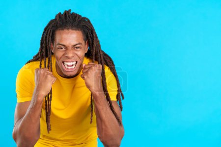 Photo for Happy hispanic man with dreadlocks gesturing with fists while celebrating in studio with blue background - Royalty Free Image