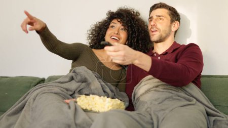 Couple switching channel on television with remote control lying on the sofa with a blanket eating popcorn