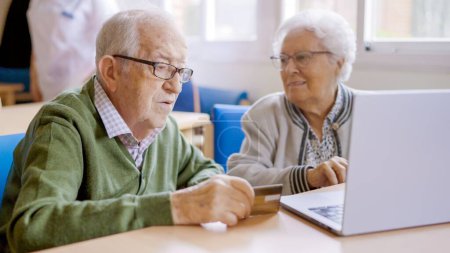 Foto de Mujer mayor explicando cómo comprar en línea a su marido usando el ordenador portátil y la tarjeta de crédito - Imagen libre de derechos