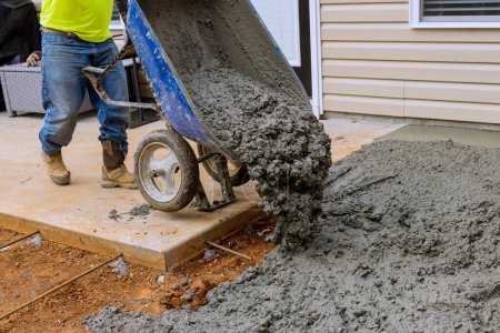 On side house construction worker pours cement from wheelbarrow to create concrete sidewalk that will be used for future