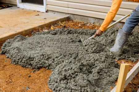 Téléchargez les photos : Travailleur de la construction verse du ciment sur le côté de la maison pour créer un nouveau trottoir qui s'étendra le long du bâtiment latéral - en image libre de droit