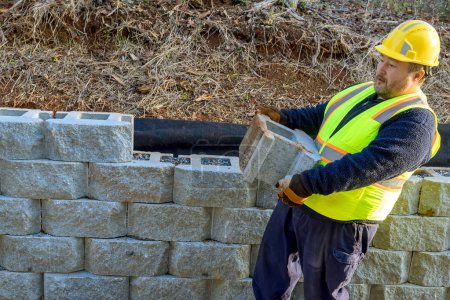 Téléchargez les photos : Travailleur de la construction levé bloc de béton avant de le monter sur le mur de soutènement. - en image libre de droit