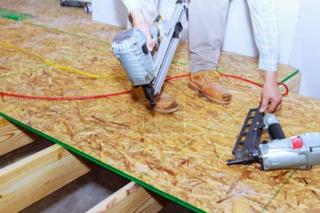 Téléchargez les photos : Nouveau plancher de la maison est installé avec contreplaqué en bois à l'aide d'un pistolet à ongles d'air - en image libre de droit