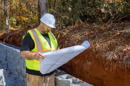 Construction worker studies used by blueprint plan when mounting retaining wall by cement blocks are being
