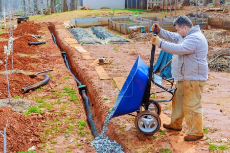 Foto de Usando carretilla, el trabajador llena las tuberías de drenaje con piedra triturada - Imagen libre de derechos