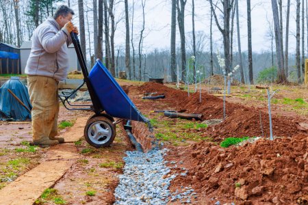 Worker uses a wheelbarrow to fill over drainage pipes with crushed stone