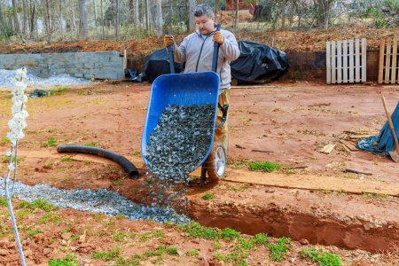 In wheelbarrow, worker fills drainage pipes with crushed stone