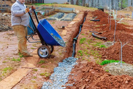 From wheelbarrow, worker fills overflowing drainage pipe with crushed stone