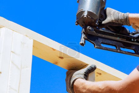 Worker in holding plank installing it on household construction using air hammer in nailing wooden beams.